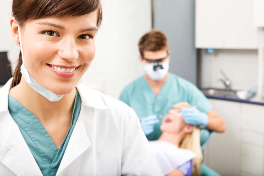 A portrait of a dental assistant smiling at the camera with the dentist working in the background